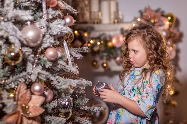 装飾されたクリスマスツリーの近くには 巻き毛の長い髪の美しい笑いの女の子が立っています クリスマスツリーを飾る クリスマスツリーに風船やおもちゃを掛けます 家庭での冬のレクリエーションの概念 閉じろ — ストック写真