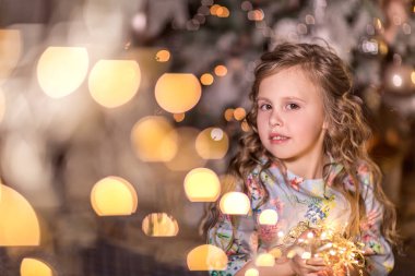 Kıvırcık uzun saçlı güzel bir kızın portresi bulanık Noel ışıkları (bokeh) ve bir Noel ağacı. Evde kış tatili kavramı. Kapat..