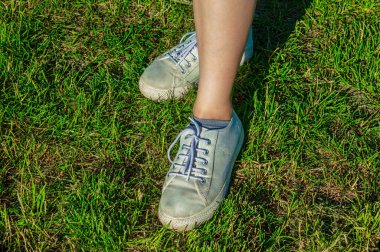 Genç bir kadın yeşil çimlerin üzerinde beyaz ayakkabılarla oturuyor. Üst Manzara. Batan güneşin uzun gölgeleri