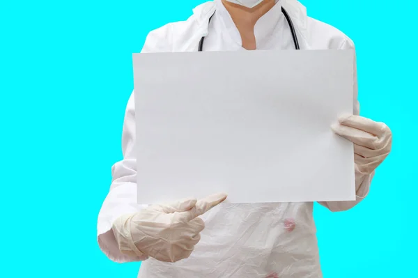 Young woman doctor in white uniform and gloves points a finger to a white blank sheet that holds in her hand. Space for text, copyspace.