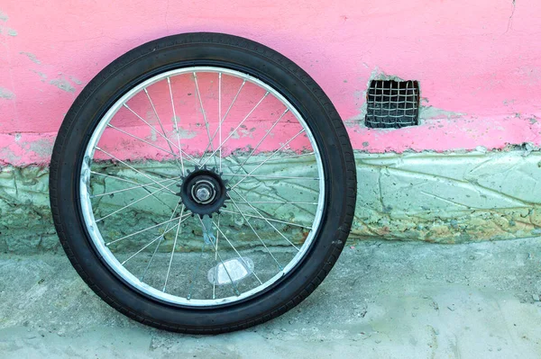 a small bicycle wheel rests on the concrete pink wall of the house