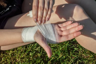 The girl is bandaging her wounded palm and wrist while sitting on the green grass. View from above. Wound care bandage, control bleeding. clipart