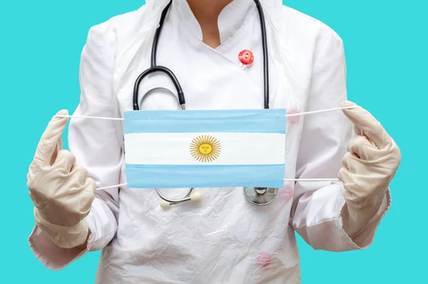 Epidemic in Argentina. Young woman doctor in a medical coat and gloves holds a medical mask with the print of the flag of Argentina on a blue background isolated