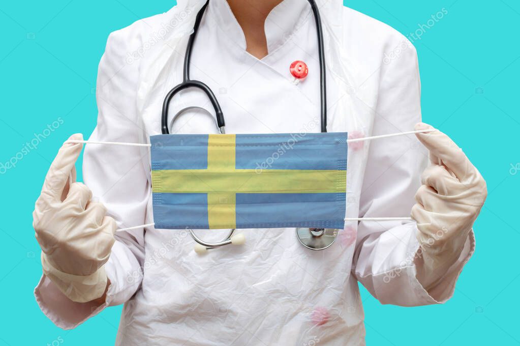 Epidemic in Sweden. Young woman doctor in a medical coat and gloves holds a medical mask with the print of the flag of Sweden on a blue background isolated.