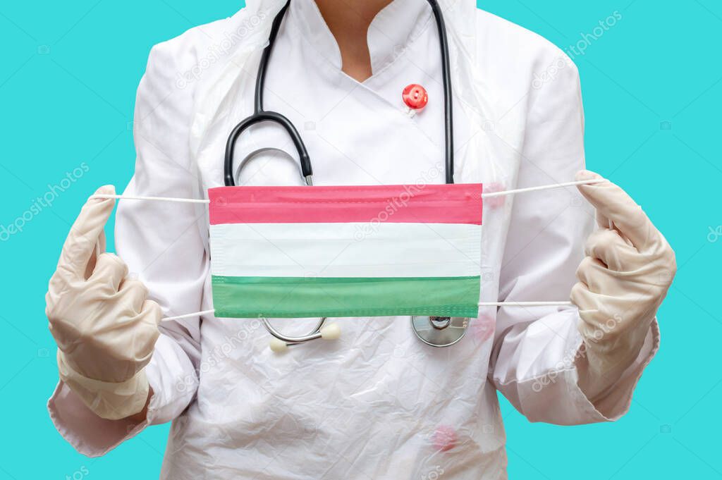 Epidemic in Hungary. Young woman doctor in a medical coat suit and gloves holds a medical mask with the print of the flag of Hungary on a blue background isolated