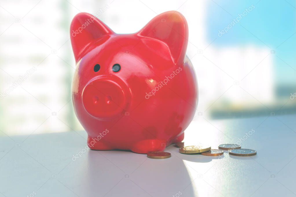 A red piggy bank stands on the table near the window, near it is a pile of small euro coins. Bright light background of city houses