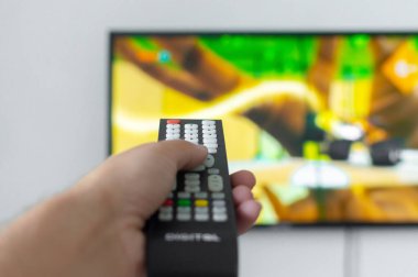Adam uzaktan kumandayı televizyona doğrulttu ve kanalı değiştirdi. Renkli bulanık TV ekranı karikatür gösteriyor. Birinci şahıs görünümü