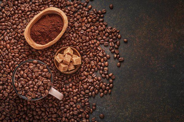 Coffee roasted beans in cup and scattered nearby, ground coffee and cane sugar on a brown table background. Top view with space to copy text.