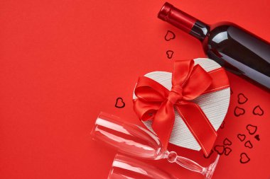 Kırmızı arka planda kırmızı kurdeleli, kalp şeklinde şarap, bardak ve hediye kutusu. Sevgililer Günü kartpostalı. Üst görünüm.