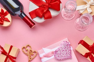 Şarap, bardak ve kalp şeklinde hediye paketi. Pembe arka planda kırmızı kurdele var. Sevgililer Günü kartpostalı. Üst görünüm.