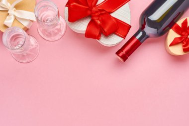 Şarap, bardak ve kalp şeklinde hediye paketi. Pembe arka planda kırmızı kurdele var. Sevgililer Günü kartpostalı. Üst görünüm.