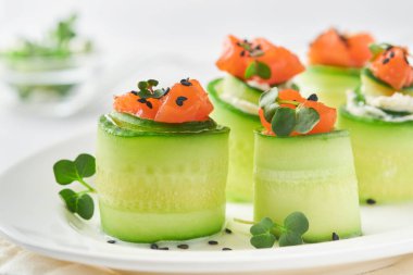 Tatil sebze aperatifleri. Salatalıklar yumuşak peynirli rulo salatalık, tuzlanmış somon, mikroyeşillik ve siyah susam, beyaz tabakta servis edilir. Seçici odak.