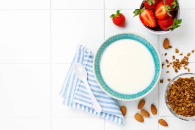 Kahvaltı gevreği ve beyaz masada taze çilek yapmak için malzemelerle dolu beyaz kasede Yunan yoğurdu. Üst görünüm.