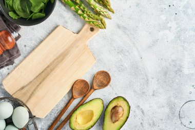 Bir demet taze yeşil organik kuşkonmaz ıspanak, avokado, tavuk yumurtası, biber sosu ve gri arka planda kesim tahtası, üst manzara. Fotokopi alanı olan yemek pişirme arka planı. Model.