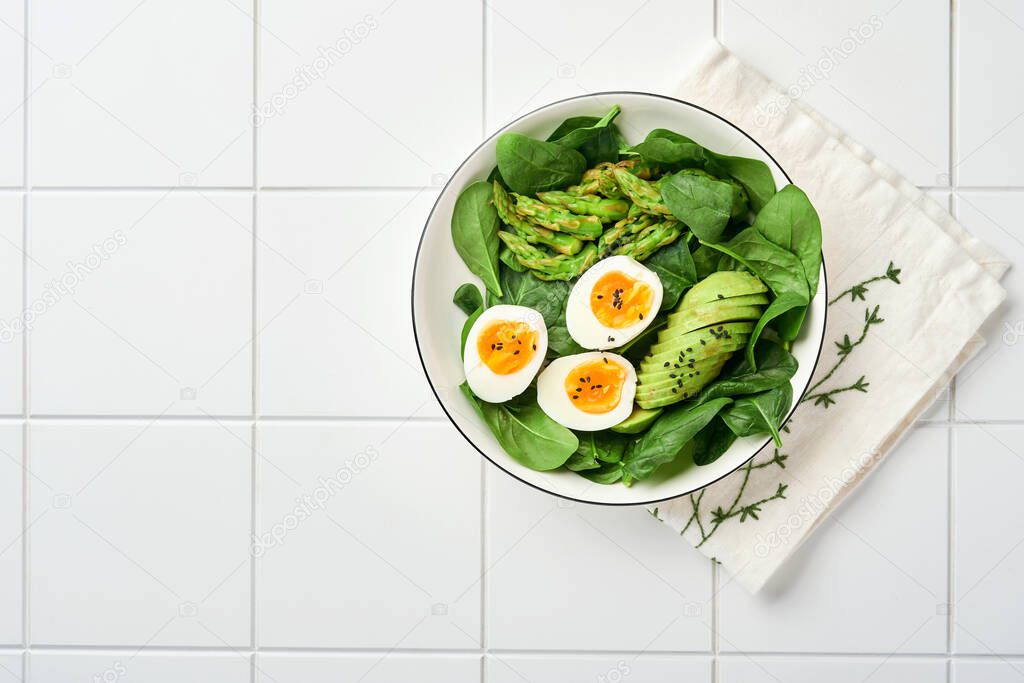 Fresh vegetable salad with avocado, asparagus, crumpled eggs with black sesame seeds and spinach on plate on light slate, stone or concrete background. Balanced lunch in bowl. Top view. Mock up.