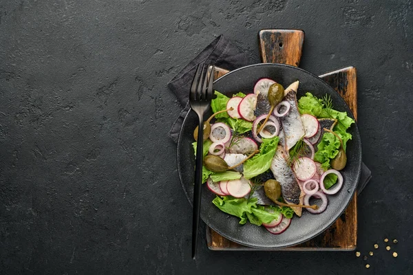 Соленая сельдь со специями, каперсами, зеленью и луковым салатом на черной тарелке на темном фоне с пространством для копирования. Маринованная нарезанная рыба. Пища со здоровыми ненасыщенными жирами, плоская — стоковое фото