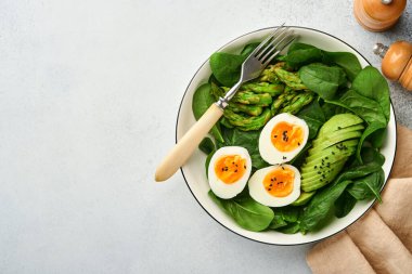 Avokadolu taze sebze salatası, kuşkonmaz, siyah susam tohumlu buruşuk yumurtalar ve ışık tabağında ıspanak, taş ya da beton arka plan. Öğle yemeğini kasede dengeledim. Üst Manzara. Model.