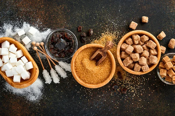 White sugar, cane sugar cubes, caramel in bamboo bowl on dark brown table concrete background. Assorted different types of sugar. Top view or flat lay.