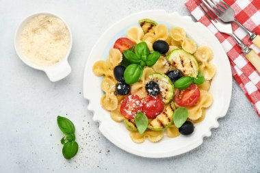 Sebzeli makarna fiocchi salatası ızgara kabak, vişneli domates, zeytin, fesleğen ve parmesan peyniri beyaz tabakta taş ya da beton arka planda. Yemek konsepti. Üst görünüm.