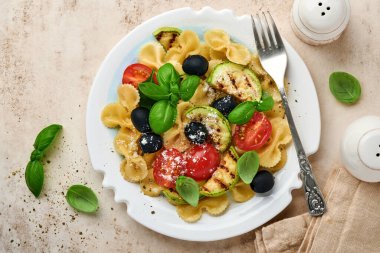 Sebzeli makarna fiocchi salatası ızgara kabak, vişneli domates, zeytin, fesleğen ve parmesan peyniri beyaz tabakta taş ya da beton arka planda. Yemek konsepti. Üst görünüm.