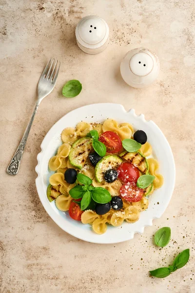 Sebzeli makarna fiocchi salatası ızgara kabak, vişneli domates, zeytin, fesleğen ve parmesan peyniri beyaz tabakta taş ya da beton arka planda. Yemek konsepti. Üst görünüm.