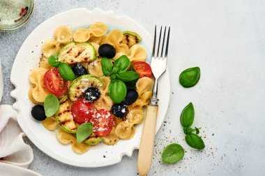Sebzeli makarna fiocchi salatası ızgara kabak, vişneli domates, zeytin, fesleğen ve parmesan peyniri beyaz tabakta taş ya da beton arka planda. Yemek konsepti. Üst görünüm.