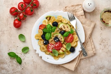 Sebzeli makarna fiocchi salatası ızgara kabak, vişneli domates, zeytin, fesleğen ve parmesan peyniri beyaz tabakta taş ya da beton arka planda. Yemek konsepti. Üst görünüm.