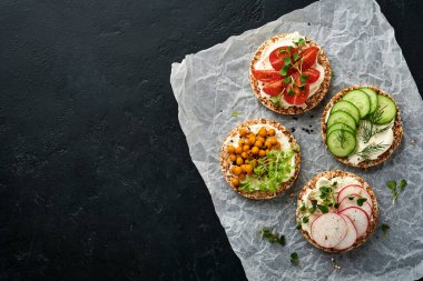 Krem peynirli, turplu, domatesli, nohutlu, salatalıklı ve mikroyeşilli dört çıtır karabuğday ekmeği siyah taş zemin üzerinde sağlıklı bir kahvaltı için. Vejetaryen ve sağlıklı beslenmeyi düşün..