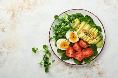 Avokado, vişneli domates, ıspanak ve tavuk yumurtası, mikro yeşil bezelyeler ve siyah susam tohumları beyaz taş masa arka planında kasede taze salata. Sağlıklı kahvaltı yemeği konsepti. Üst görünüm.