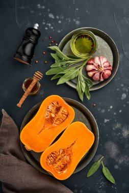 Yarım parça çiğ organik tereyağlı kabak, adaçayı yaprağı, çok renkli sarımsak, bal, siyah tahtada tuz ve biber, taş ya da beton arka plan. Yemek arkaplanı. Kopyalama alanı olan üst görünüm.