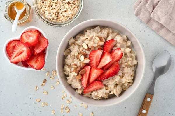 stock image Oatmeal porridge with strawberry slices, nuts almonds and honey in bowl on grey table. Healthy eating, dieting, vegetarian food concept. Place for text