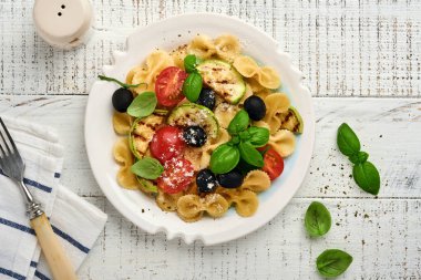 Sebzeli makarna fiocchi salatası ızgara kabak, vişneli domates, zeytin, fesleğen ve parmesan peyniri beyaz tabakta taş ya da beton arka planda. Yemek konsepti. Üst görünüm