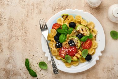 Sebzeli makarna fiocchi salatası ızgara kabak, vişneli domates, zeytin, fesleğen ve parmesan peyniri beyaz tabakta taş ya da beton arka planda. Yemek konsepti. Üst görünüm