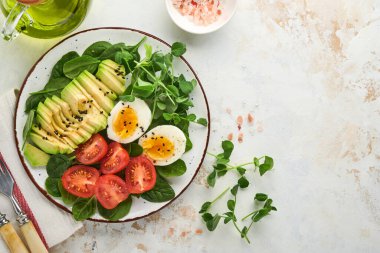 Avokado, vişneli domates, ıspanak ve tavuk yumurtası, mikro yeşil bezelyeler ve siyah susam tohumları beyaz taş masa arka planında kasede taze salata. Sağlıklı kahvaltı yemeği konsepti. Üst görünüm.