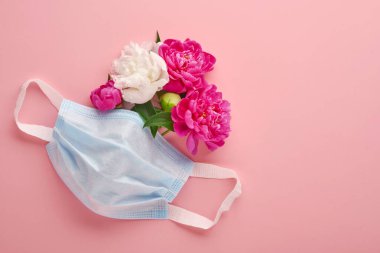 Koruyucu tıbbi yüz maskesi, şakayık çiçekleri ve pastel pembe arka plan. Coronavirus ya da covid-19 pandemik koruma sembolü olarak tek kullanımlık cerrahi maske. Valentine, Women or Mother Day konsepti.