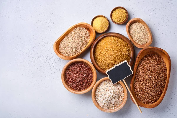 White, brown and red rice, buckwheat, millet, corn groats, quinoa and bulgur in wooden bowls on the light gray kitchen table. Gluten-free cereals. Top view with copyspace