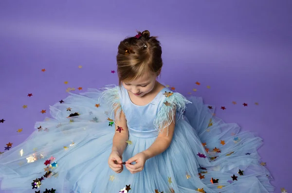 Kleines Mädchen im Prinzessinnenkleid spielt mit Konfetti auf lila Hintergrund — Stockfoto