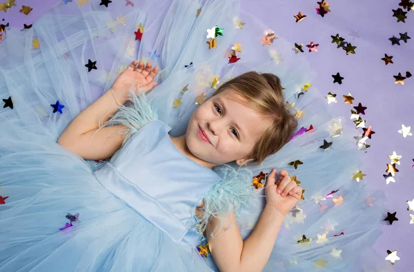 En liten flicka ligger på golvet med konfetti och tittar på kameran på en lila bakgrund med utrymme för text — Stockfoto