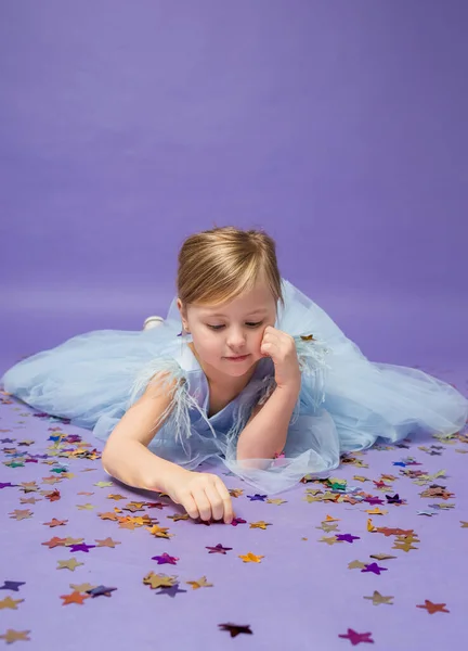 Küçük bir kız yerde yatar ve mor arka planda konfetilerle oynar.