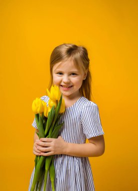 Çizgili elbiseli mutlu küçük bir kız sarı arka planda sarı laleler tutuyor.