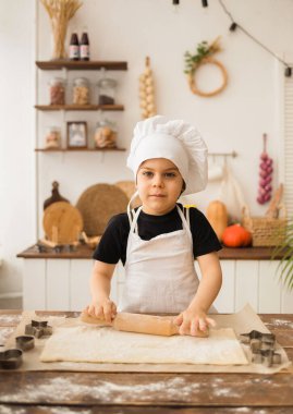 Küçük bir çocuk kepli bir yemek pişiriyor ve önlük mutfakta merdaneyle hamuru yuvarlıyor ve kameraya bakıyor.