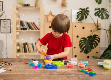 Kırmızı tişörtlü küçük bir çocuk odadaki tahta bir masada plasticine ile oynuyor. İnce motor becerilerin gelişimi.
