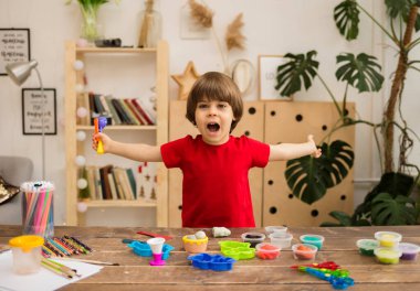 Kırmızı tişörtlü mutlu çocuk odadaki tahta bir masada plastik ve kalıplarla oynuyor. İnce motor becerilerin gelişimi