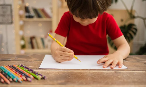 Küçük çocuk kırmızı tişörtlü bir çocuk masaya oturur ve beyaz kağıda kurşun kalemle çizer.