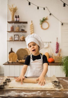 Takım elbiseli neşeli küçük şef ahşap bir masaya oturur ve mutfakta hamur toplar.