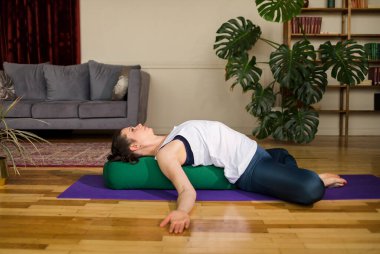 Spor kıyafetli esmer kadın yoga pozu veriyor. Odadaki paspasın üstünde destekleyici ile.