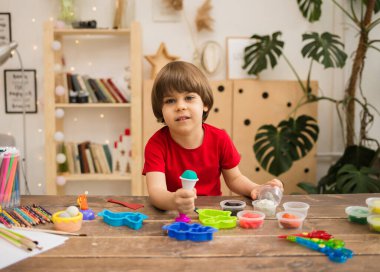 Kırmızı tişörtlü neşeli küçük çocuk, ofis malzemeleriyle tahta bir masada plasticinle oynuyor. Çocuğun yaratıcı gelişimi