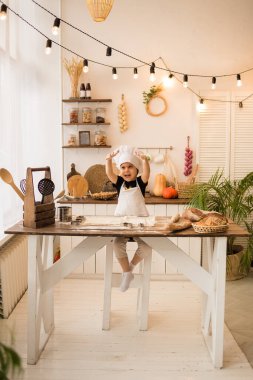 Beyaz şapkalı ve önlüklü küçük şef mutfak masasında hamur ve unla oynuyor.