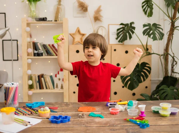 Kırmızı tişörtlü mutlu çocuk odada plastik ve plastik kalıplı bir masada oturuyor.