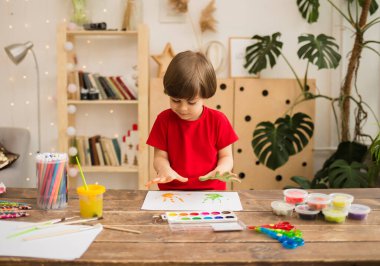 Hevesli küçük çocuk odadaki bir masada ellerini beyaz kağıda koyup resim çiziyor. Erken sanatsal gelişim